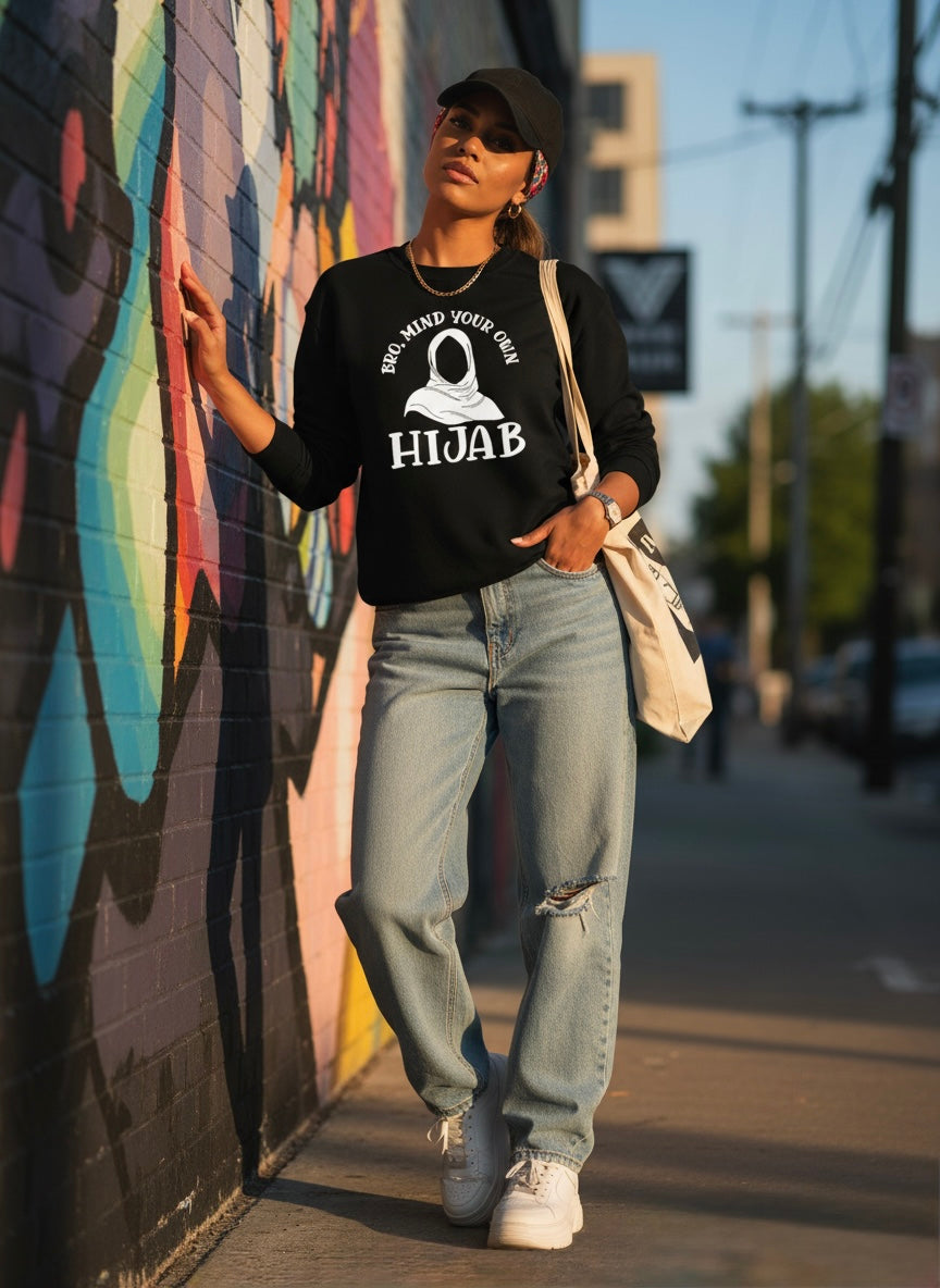 Person wearing a black hoodie with hijab text, light blue jeans, and white sneakers standing against a colorful mural.