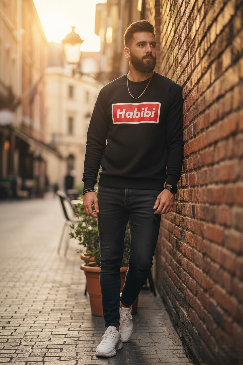 Man wearing a black sweatshirt with 'Habibi' logo standing against a brick wall on a street.