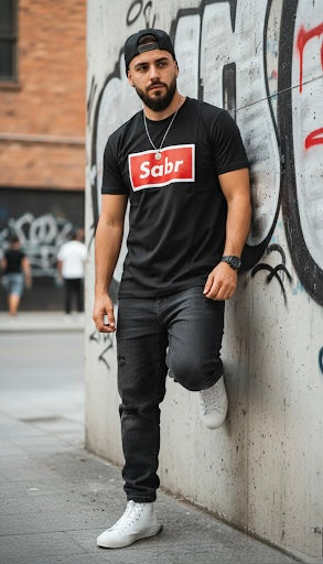 Man wearing a black t-shirt with 'Sabr' logo, black cap, and white sneakers leaning against a graffiti-covered wall.