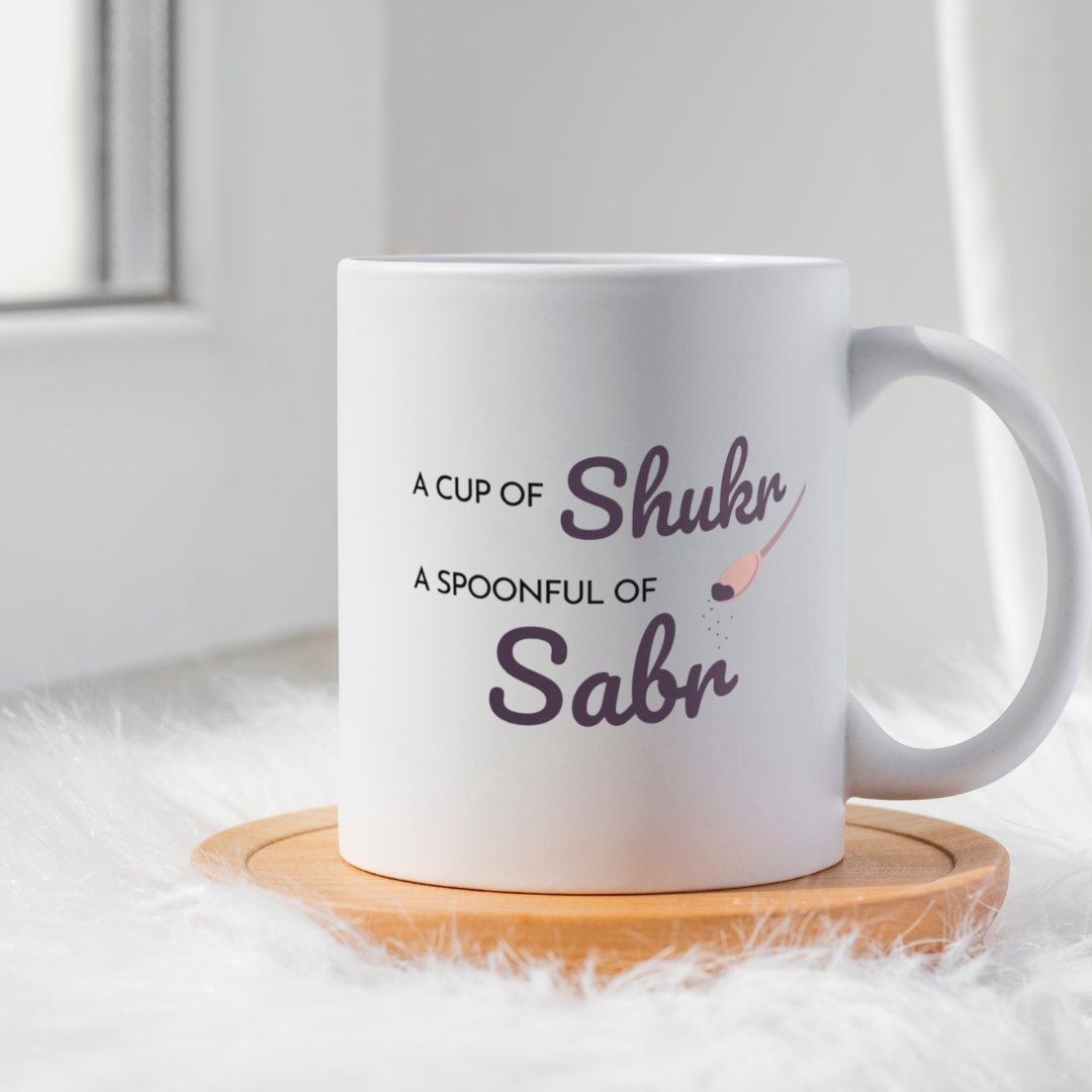 White ceramic mug with plum text saying A Cup of Shukr A Spoonful of Sabr with a spoon illustration.