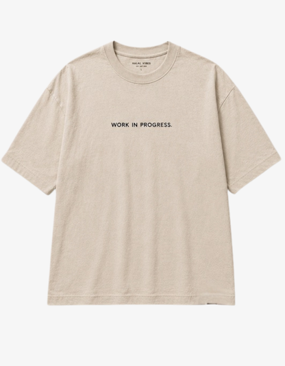 A light beige crewneck t-shirt with 'WORK IN PROGRESS.' printed in black capital letters across the chest, displayed flat against a light gray background.