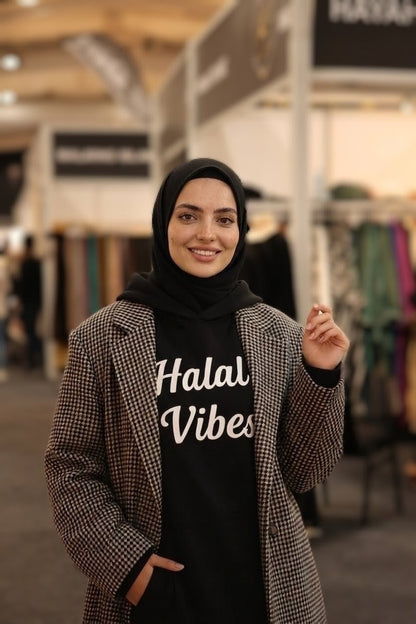 Woman wearing a black 'Halal Vibes' hoodie with a checkered coat in an indoor setting.