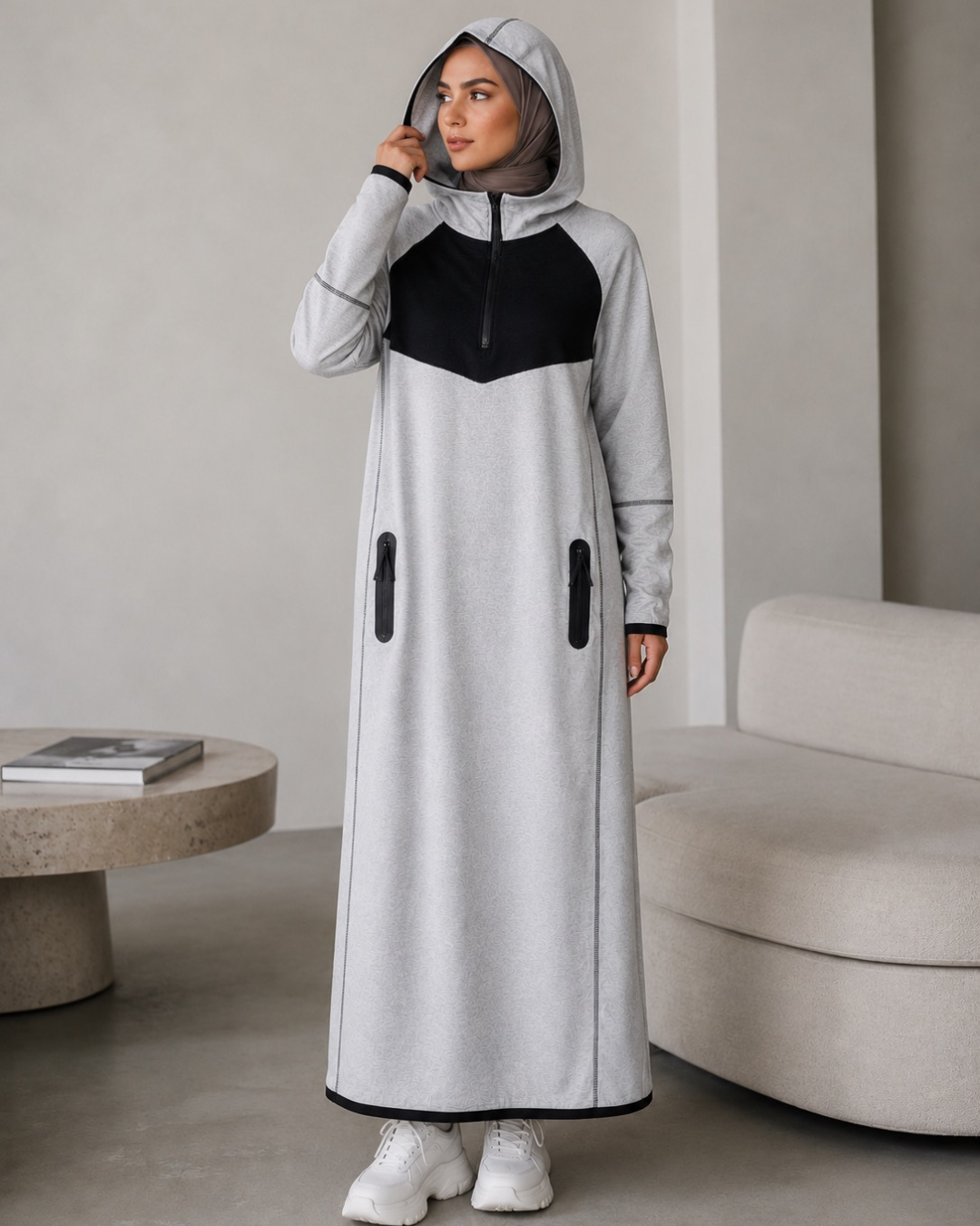 Person wearing a gray and black long dress with a hood in a minimalistic room.