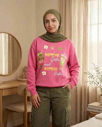 Person wearing a pink sweatshirt with text and graphics, standing in a room with a mirror and desk.